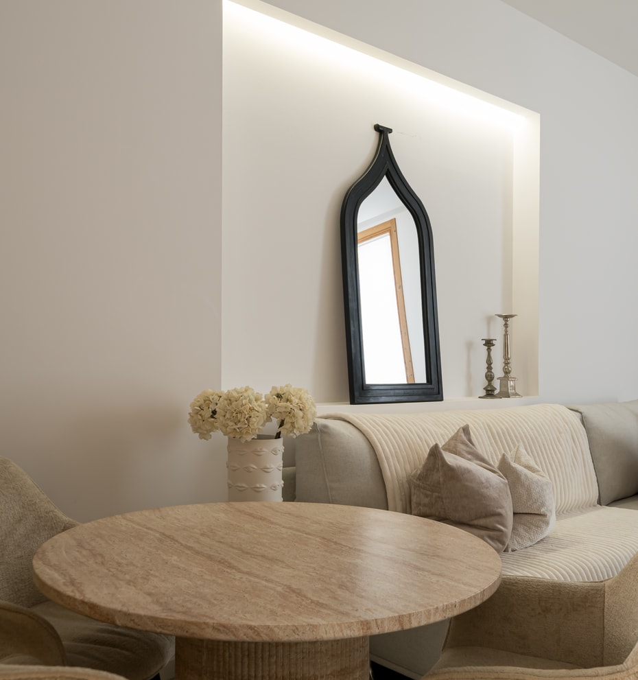 A round travertine stone dining table set in a living room with a beige textured sofa and a black arched mirror on the wall.