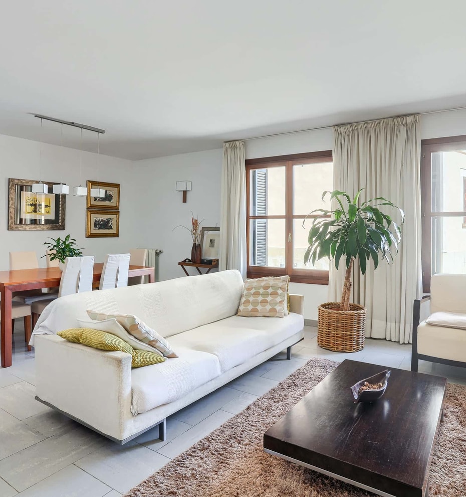 A bright living room with a white sofa, dark wooden coffee table, and a dining table for six, featuring neutral decor.