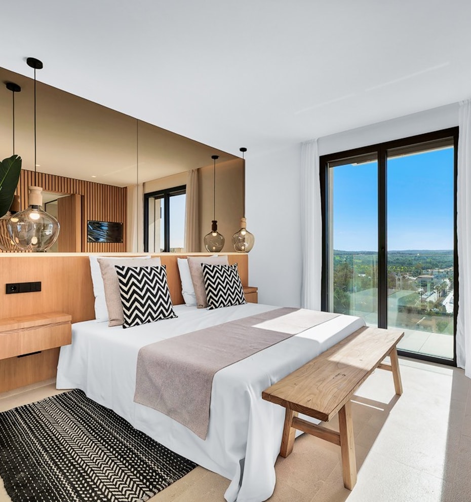 A modern bedroom featuring a bed with a wooden headboard, a large mirror, a wooden bench, and a floor-to-ceiling glass door.