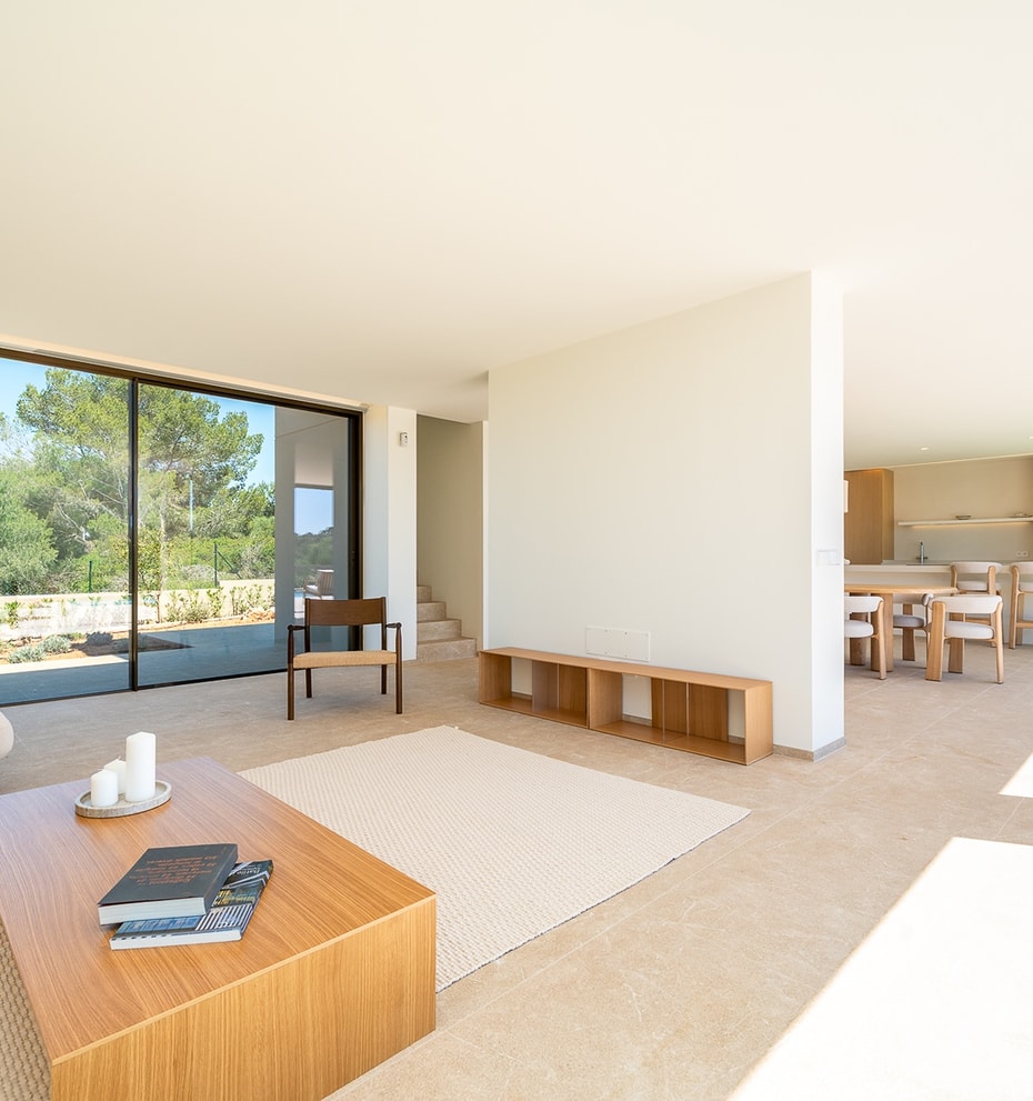A bright, open-plan living room with a light beige sofa, wooden coffee table, and large sliding glass doors overlooking trees.
