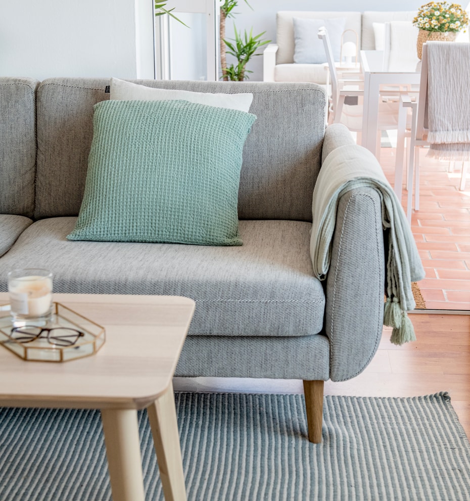 Close-up of a grey woven sofa with a mint green cushion and a light wood coffee table on a striped rug, leading to a terrace.