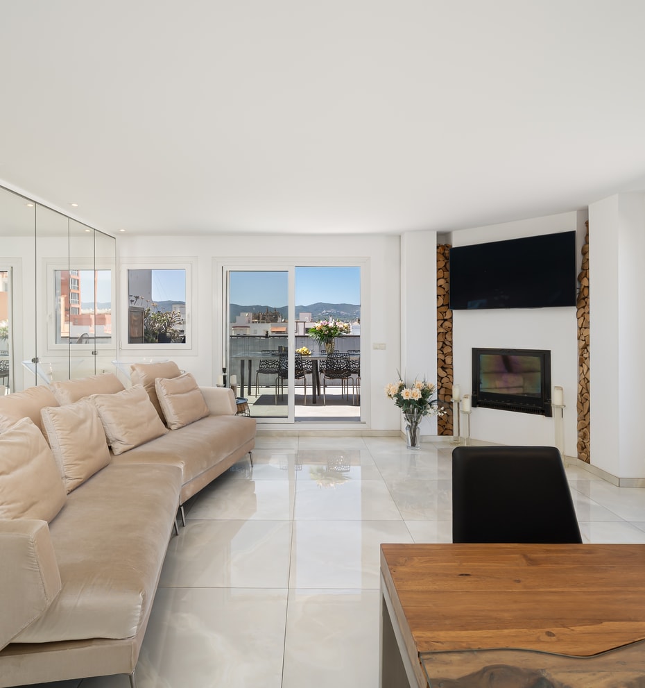 A bright, spacious living room with polished white floors, a long beige sofa, floor-to-ceiling mirrored walls, and a contemporary fireplace.