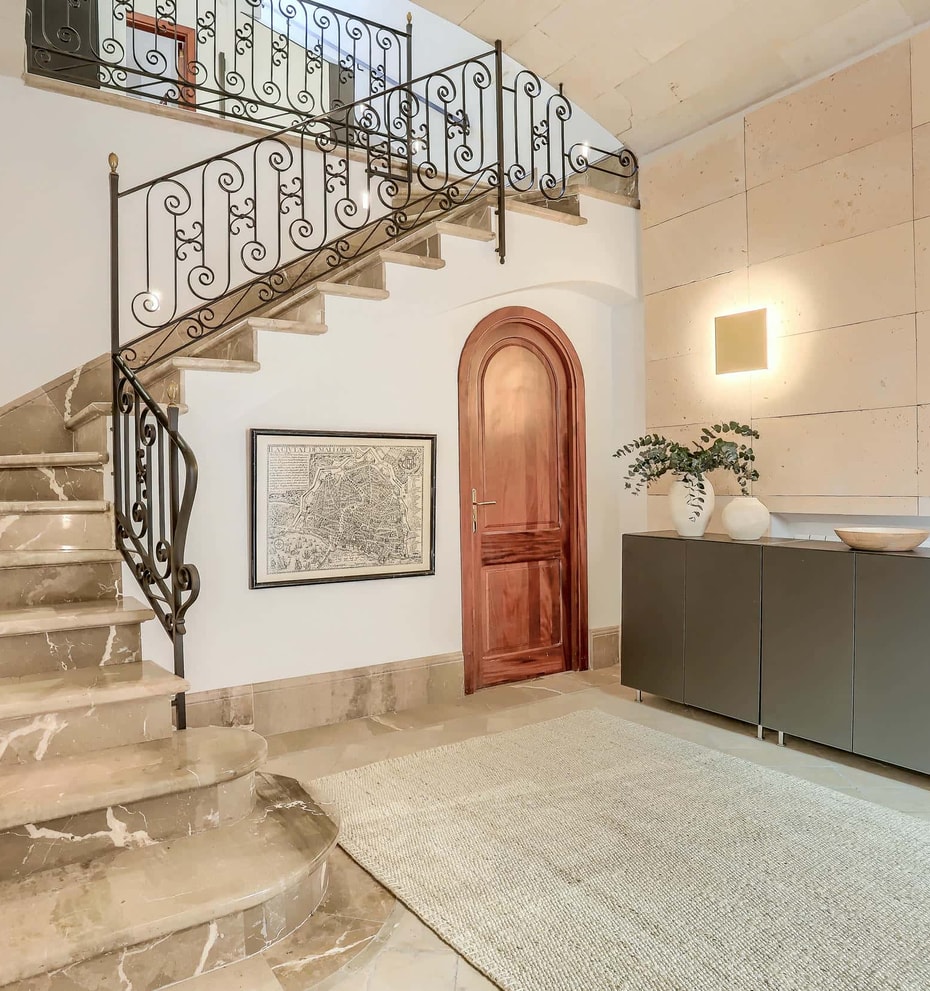 An interior shot of an entrance hall featuring a wide marble staircase with a wrought-iron railing, a curved wooden door, and a textured stone feature wall.