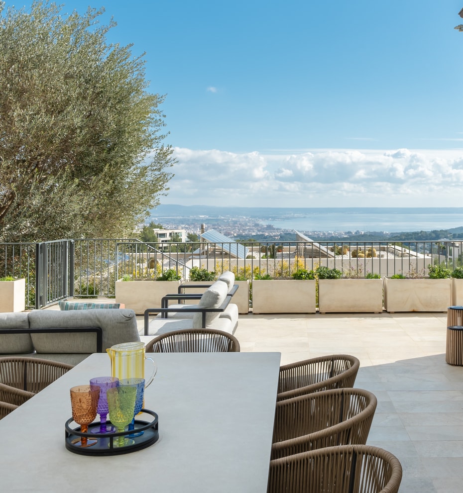 Outdoor dining terrace featuring a stone table, woven chairs, and a large olive tree overlooking the Son Vida landscape.