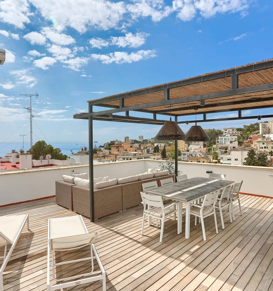 A spacious outdoor rooftop terrace featuring a wooden deck, a covered dining area with a woven pergola, lounge seating, and distant sea views over a coastal neighborhood.