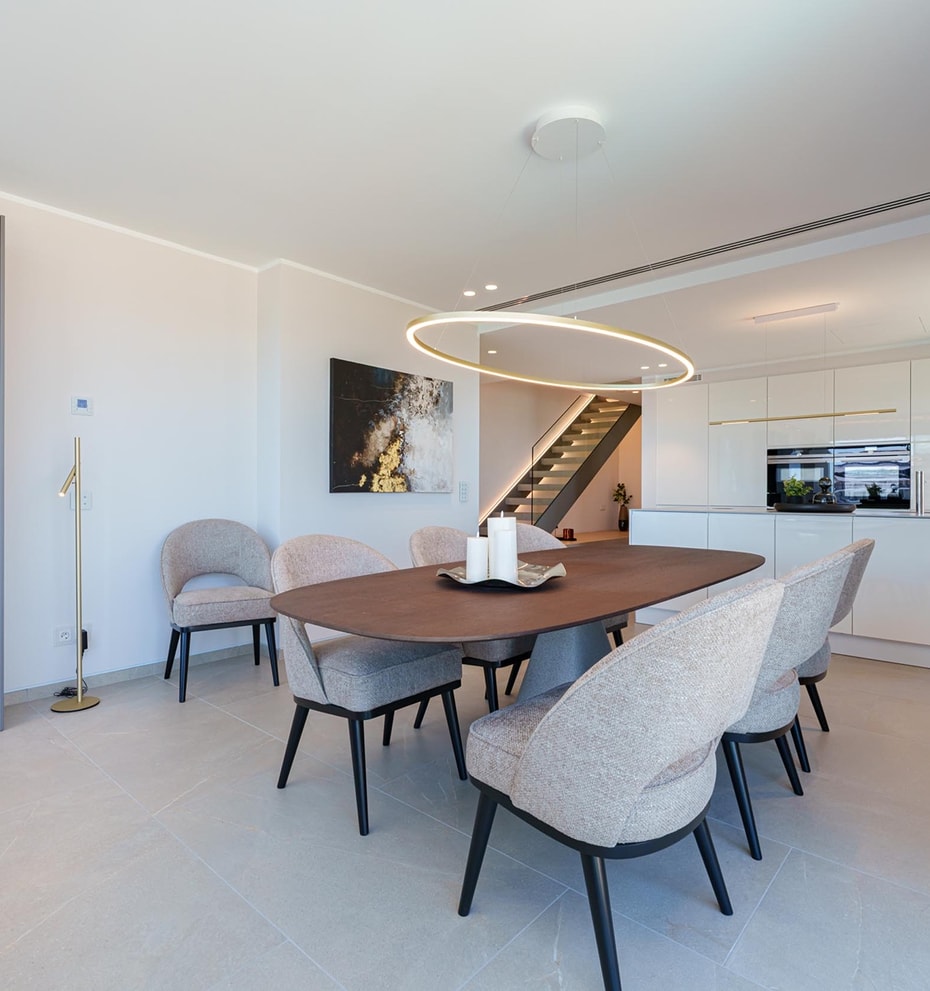 Comedor con mesa ovalada de madera y seis sillas, junto a una cocina blanca minimalista con iluminación empotrada.