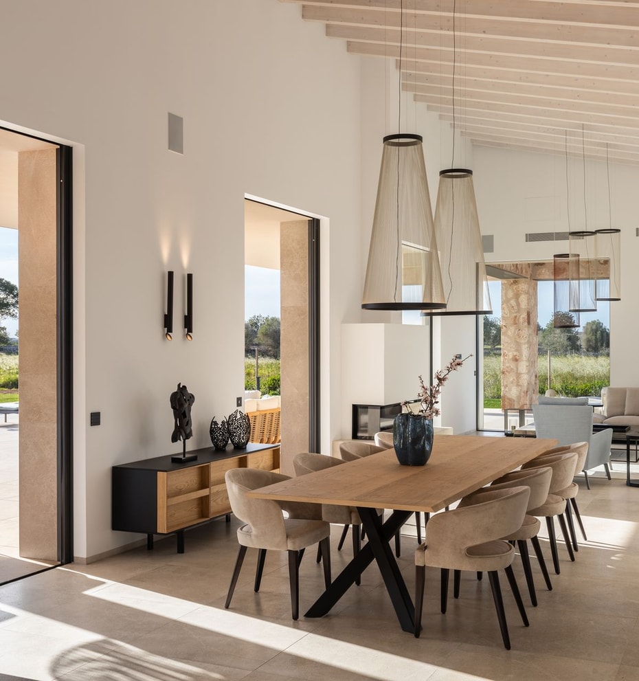 Dining room with a long oak table and cushioned chairs, open to an outdoor terrace, with white walls and exposed ceiling beams.