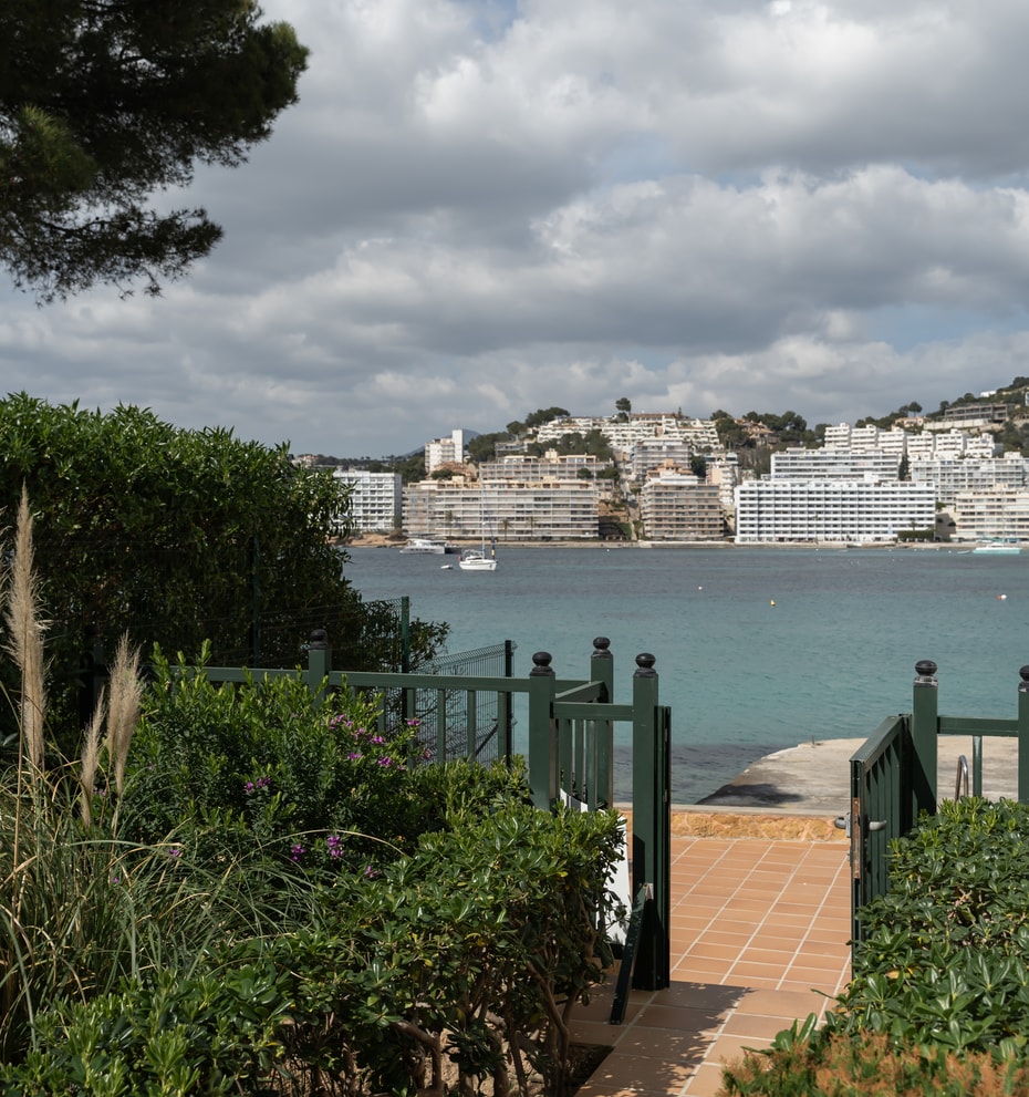 A view through a dark green garden gate over a terracotta terrace toward a calm turquoise bay and coastal buildings.