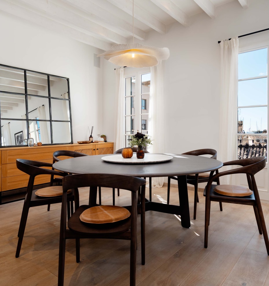 Round dark wood dining table with woven-seat chairs beneath a white beamed ceiling, French doors opening to rooftop views.