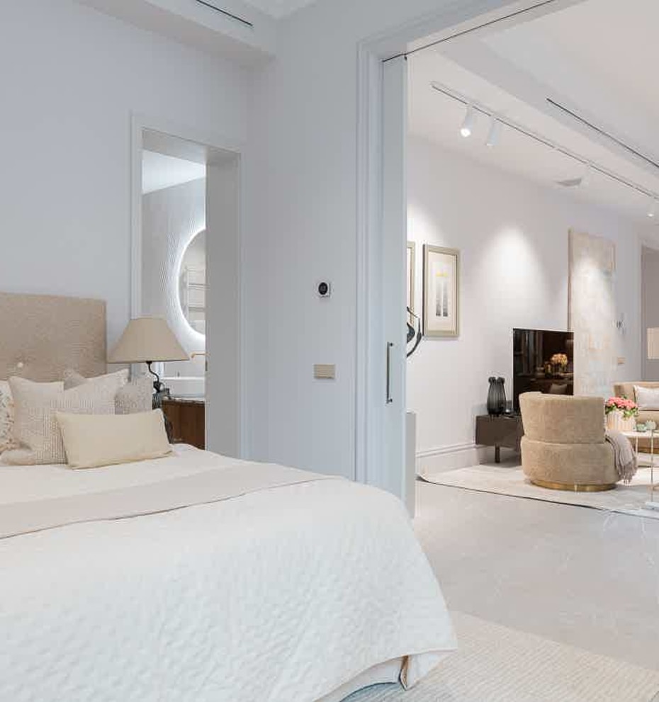 Interior view from a bedroom into an open-plan living room, featuring a beige headboard and neutral white decor.