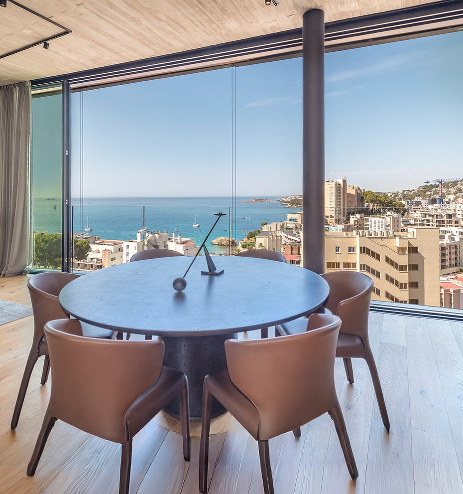 Round dark wood dining table surrounded by leather chairs next to floor-to-ceiling windows overlooking the sea and coastline.