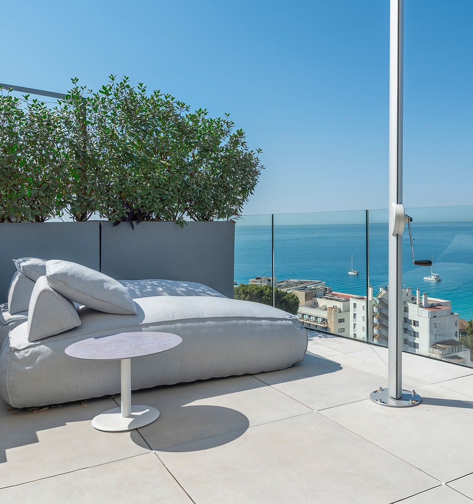 A rooftop terrace featuring a large gray outdoor daybed, a white side table, and a glass balustrade overlooking the sea.