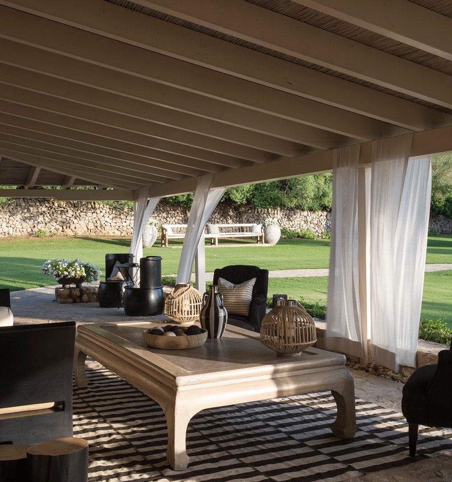 A shaded outdoor lounge area under a wooden pergola, featuring dark upholstered furniture, a central wooden coffee table, and white flowing curtains, overlooking a manicured lawn and stone boundary wall.