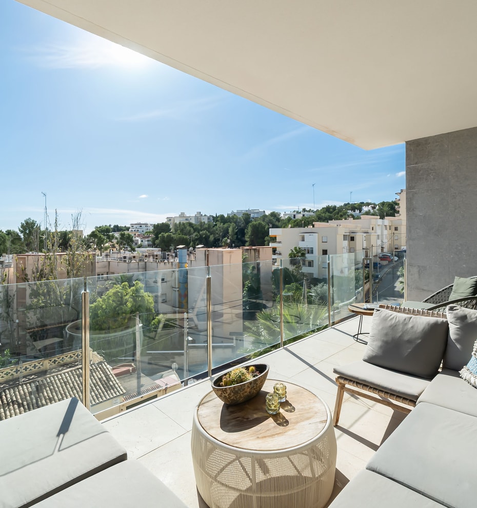 A covered terrace with grey outdoor sofas, a circular wooden coffee table, and glass railings overlooking a coastal town.