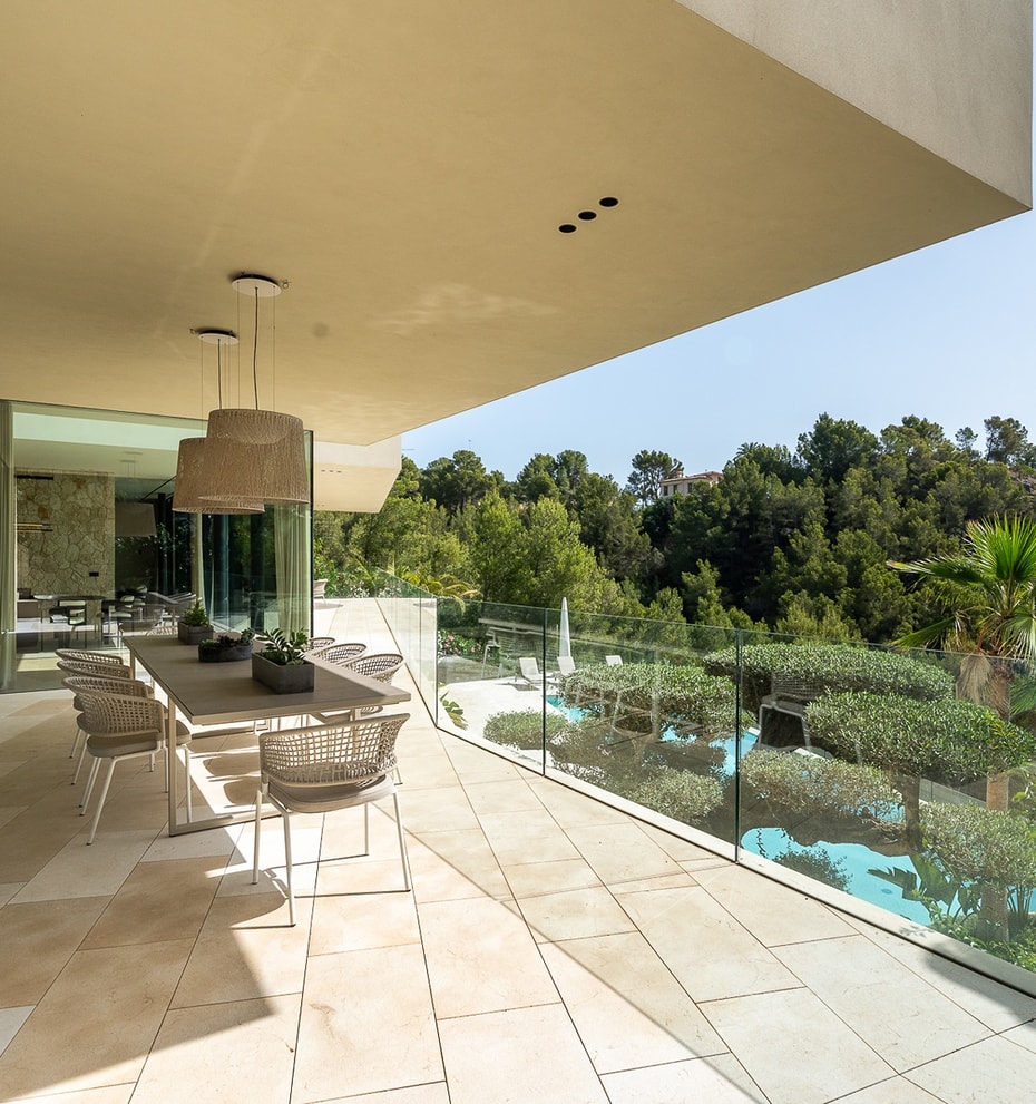 An expansive covered outdoor terrace featuring a dining table, natural stone flooring, and a glass balustrade overlooking trees.