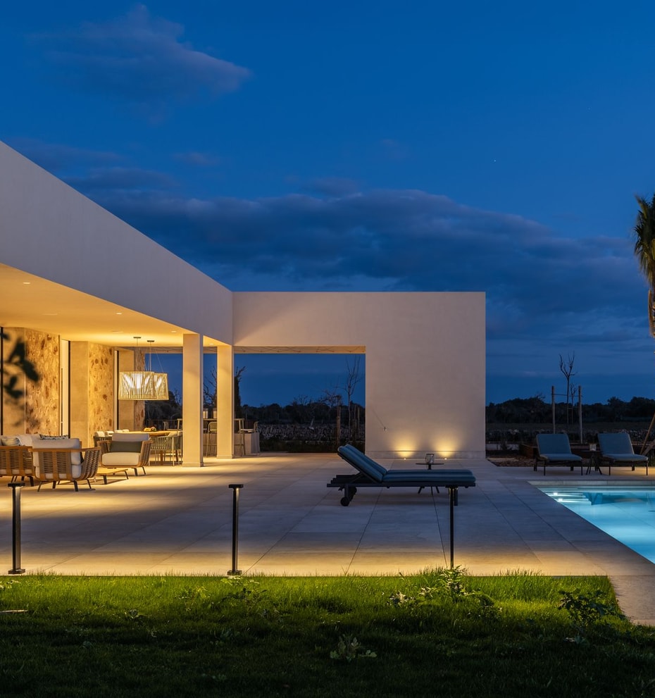 Exterior view of a modern finca at twilight, featuring a lit swimming pool, stone terrace, and warm architectural lighting.