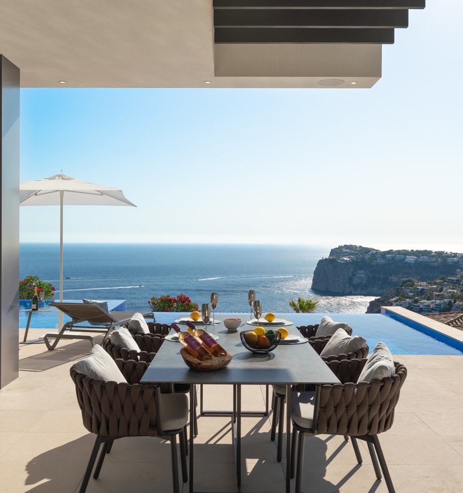 Covered outdoor dining terrace with a dark metal table, woven chairs, and an infinity pool edge against a Mediterranean sea view.