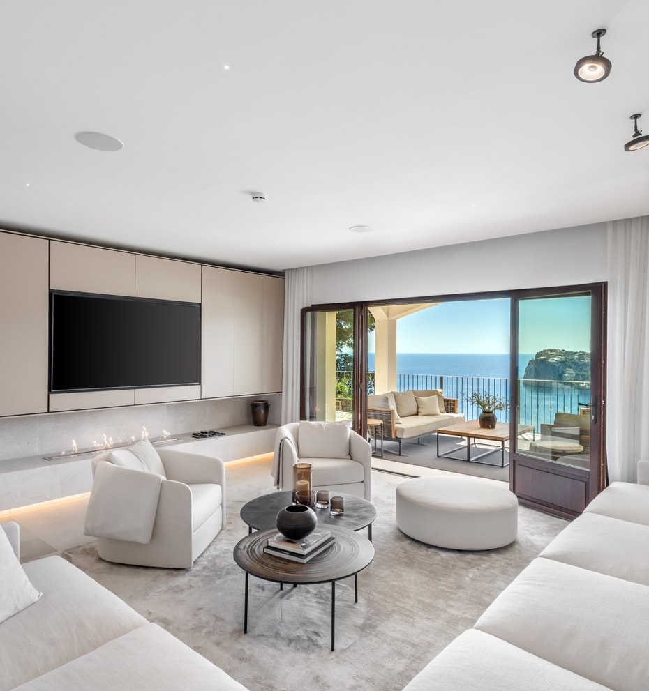Modern white living room with soft seating and circular tables facing wide glass doors and a coastal view.