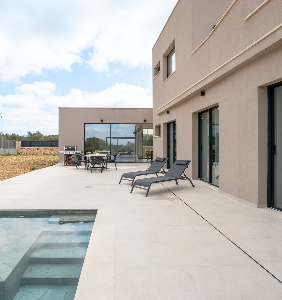 Wide view of a modern villa terrace featuring a rectangular swimming pool, sun loungers, and large sliding glass doors.