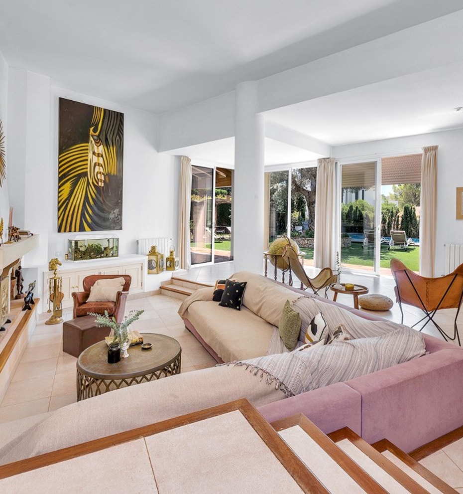 A bright, open-plan living room with a large corner sofa, a fireplace, and glass doors leading to an outdoor garden area.
