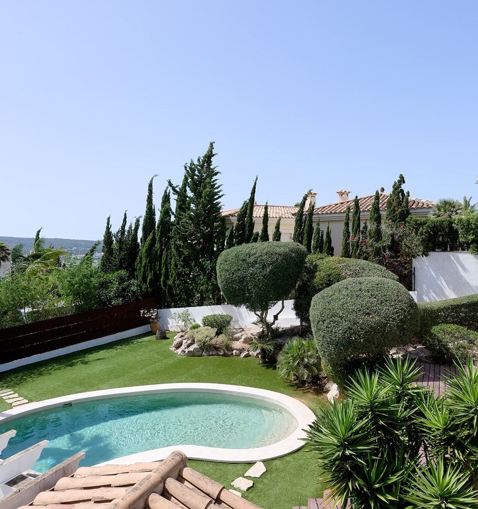 Vista desde tarima de madera sobre piscina ovalada, césped artificial, topiaria y palmeras con montañas al fondo
