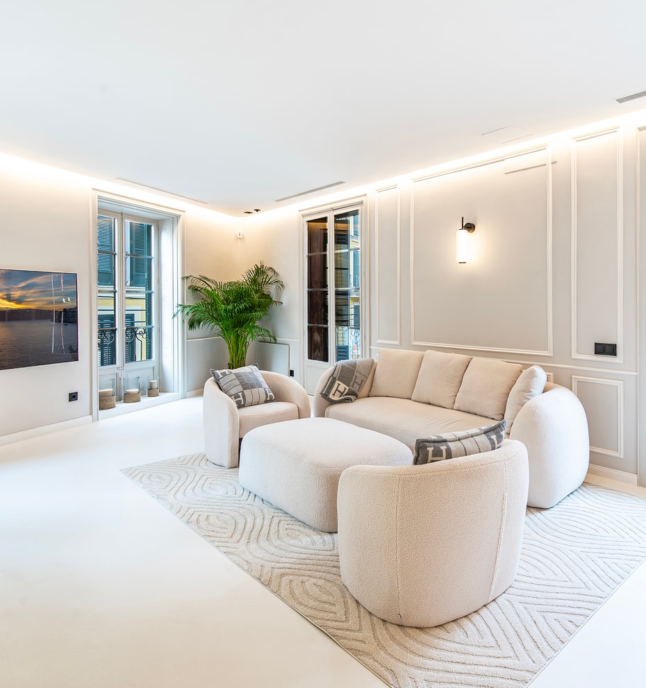 A bright, modern living room featuring cream-colored textured armchairs, a large sofa, and white walls with vertical paneling.