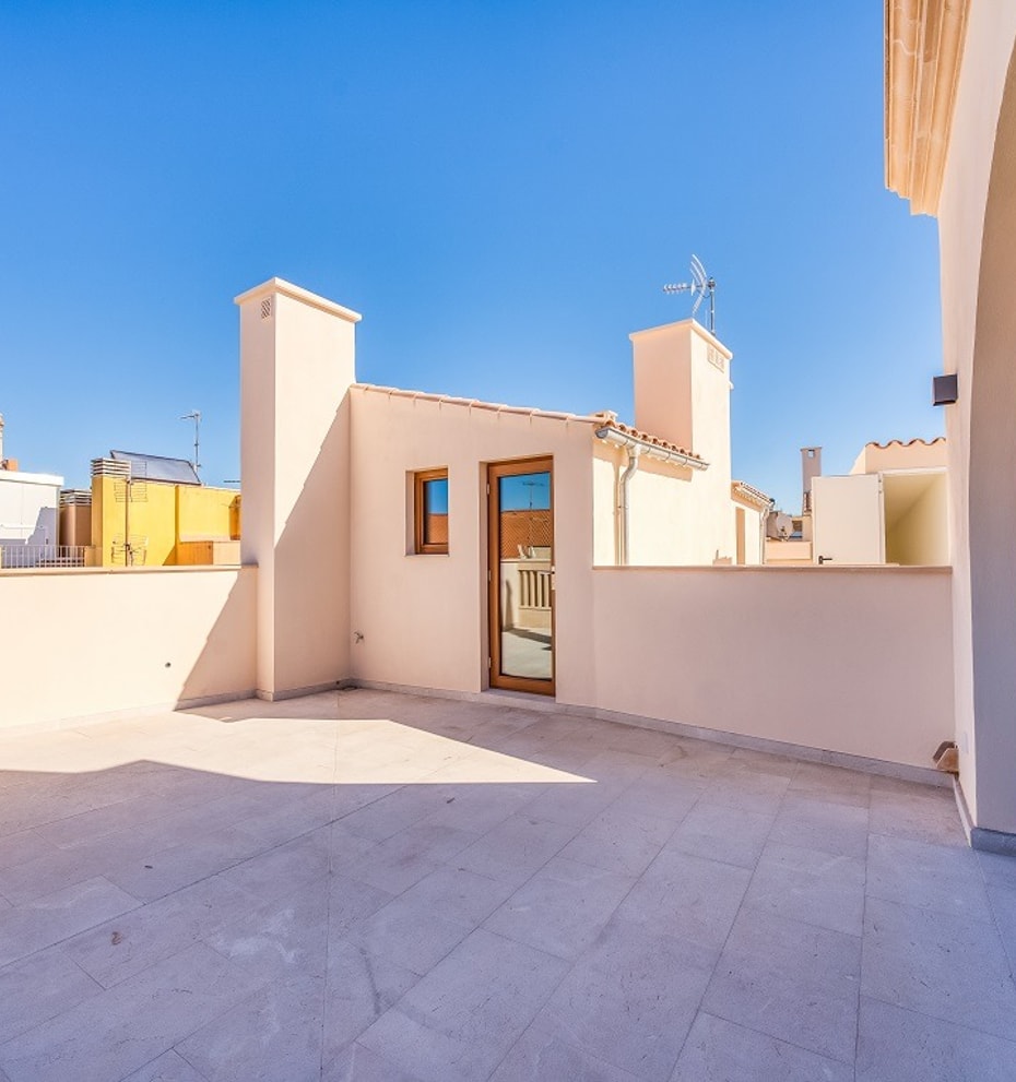 An open stone-paved rooftop terrace with light-colored walls and glass doors under a clear blue sky, overlooking city buildings.