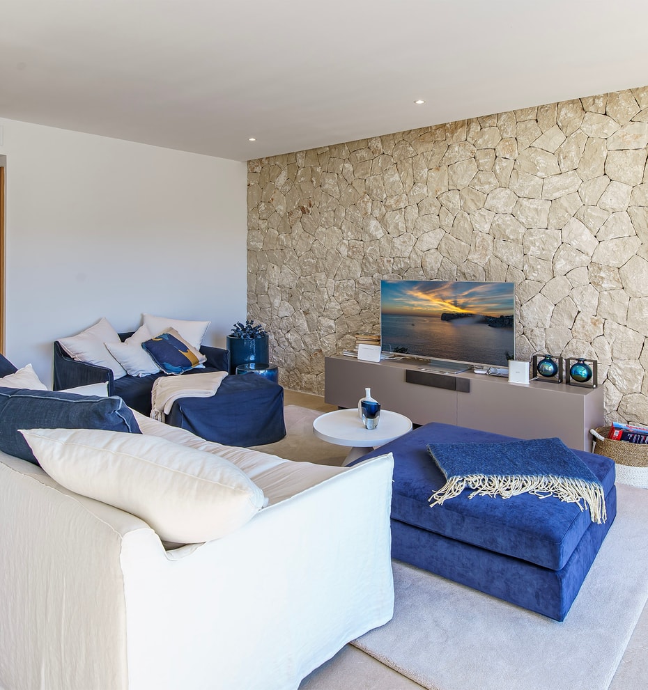 Modern living room featuring a textured natural stone wall, white sofa, blue armchairs, and neutral tiled flooring.
