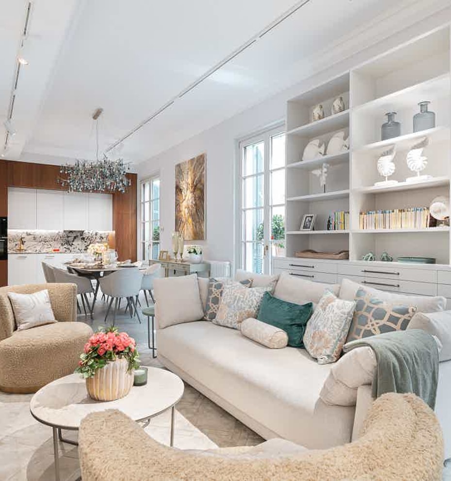 Open-plan living room with white custom built-in shelving, a neutral beige sofa, and two plush armchairs facing a kitchen area.