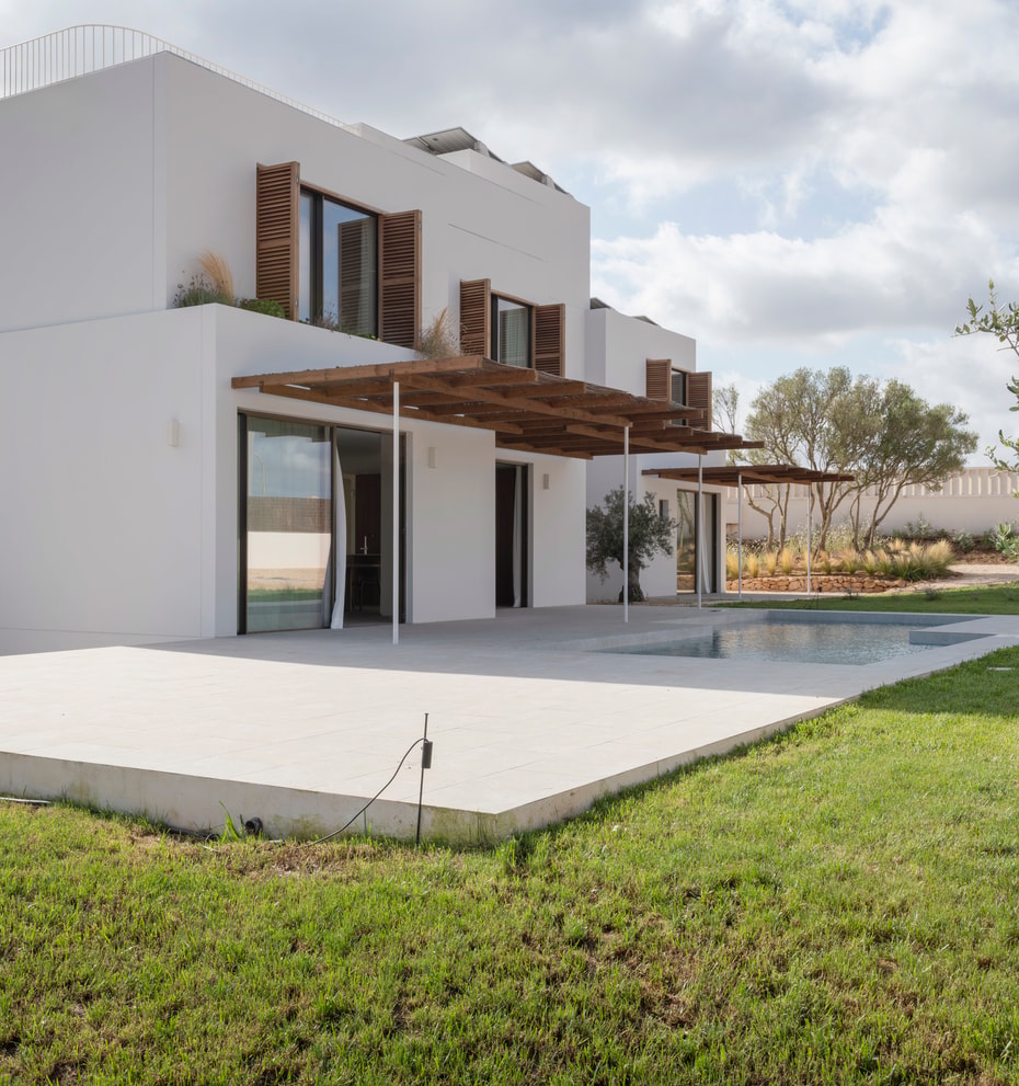 Modern two-story white villa with wooden shutters, a large pergola, and stone terrace overlooking a green lawn.