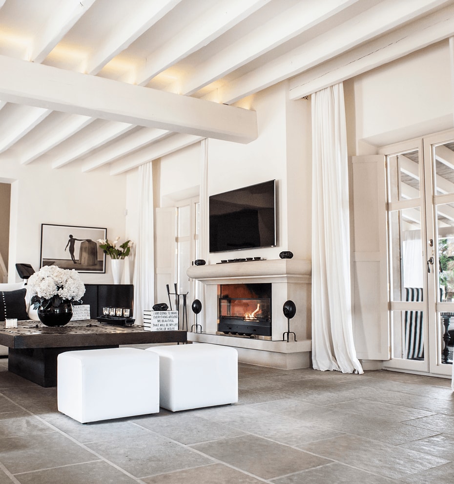Bright living area with a stone fireplace, exposed beam ceiling, stone flooring, and white linen curtains.