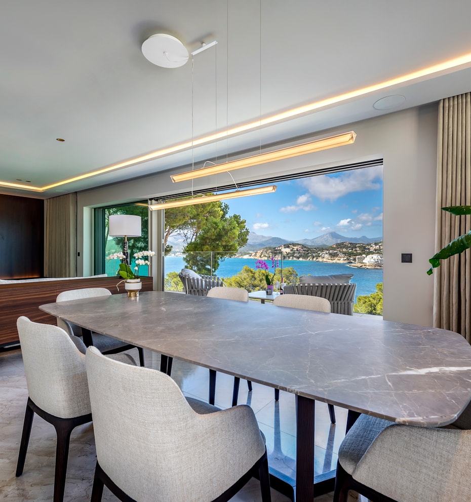 Dining room with a large oval grey stone table and upholstered chairs, facing an open terrace overlooking a blue bay.
