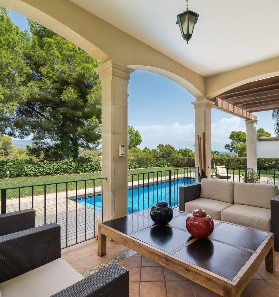 A covered terrace with comfortable outdoor seating overlooks a blue swimming pool and surrounding green gardens on a sunny day.