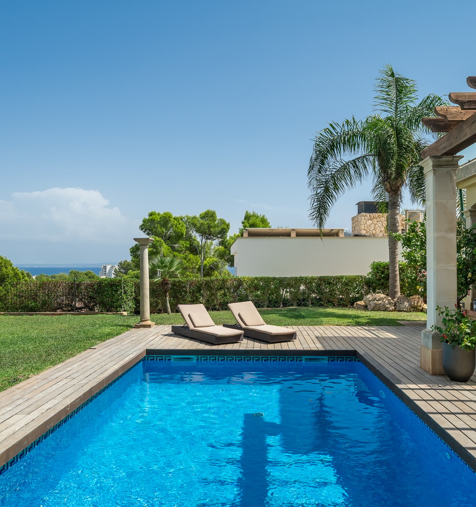 Rectangular swimming pool with blue water next to a timber deck, two sun loungers, and a shaded pergola in a sunny garden.