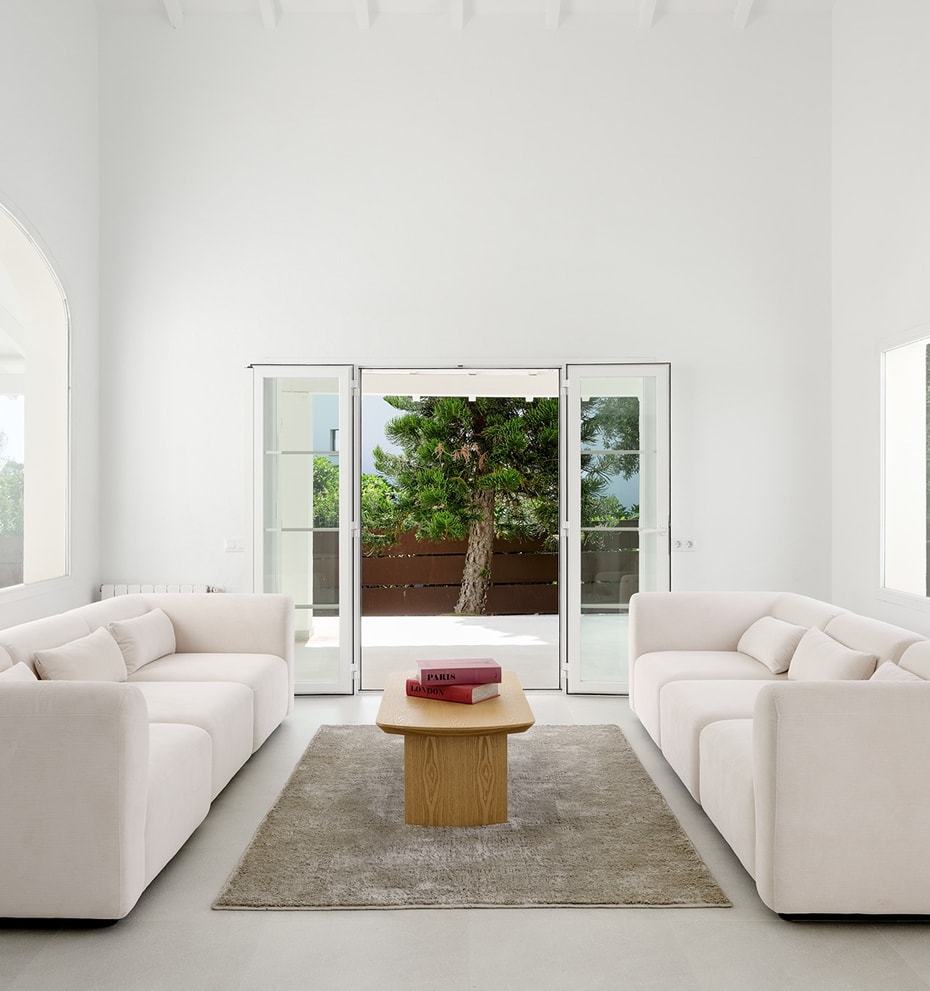 Salón blanco de doble altura con ventana arqueada, dos sofás color crema y puertas acristaladas hacia el jardín