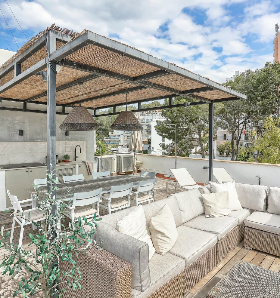 A rooftop terrace featuring a wicker sectional sofa, dining area, and outdoor kitchen under a wooden pergola.