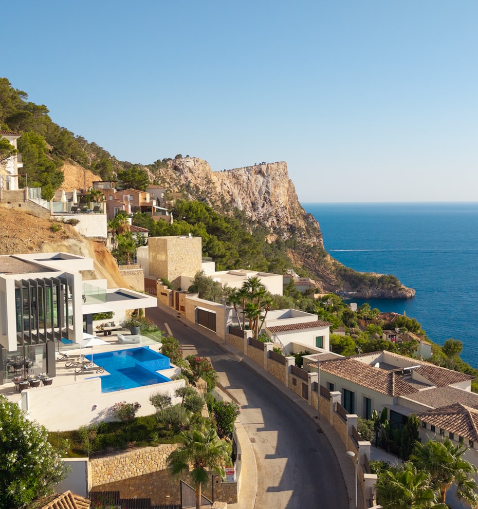 Aerial view of a white cliffside villa with a blue swimming pool, situated along a coastal road overlooking the Mediterranean.