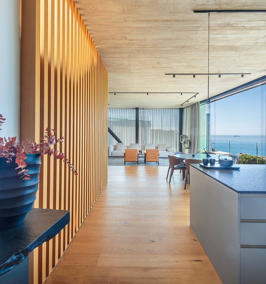 Interior of a modern kitchen showing a textured wood slat wall divider, dark kitchen island, and open-plan living area with horizon sea views.