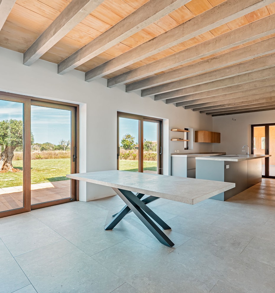 Dining area with a stone table and dark metal legs, exposed wooden ceiling beams, and large glass doors overlooking a rural landscape.