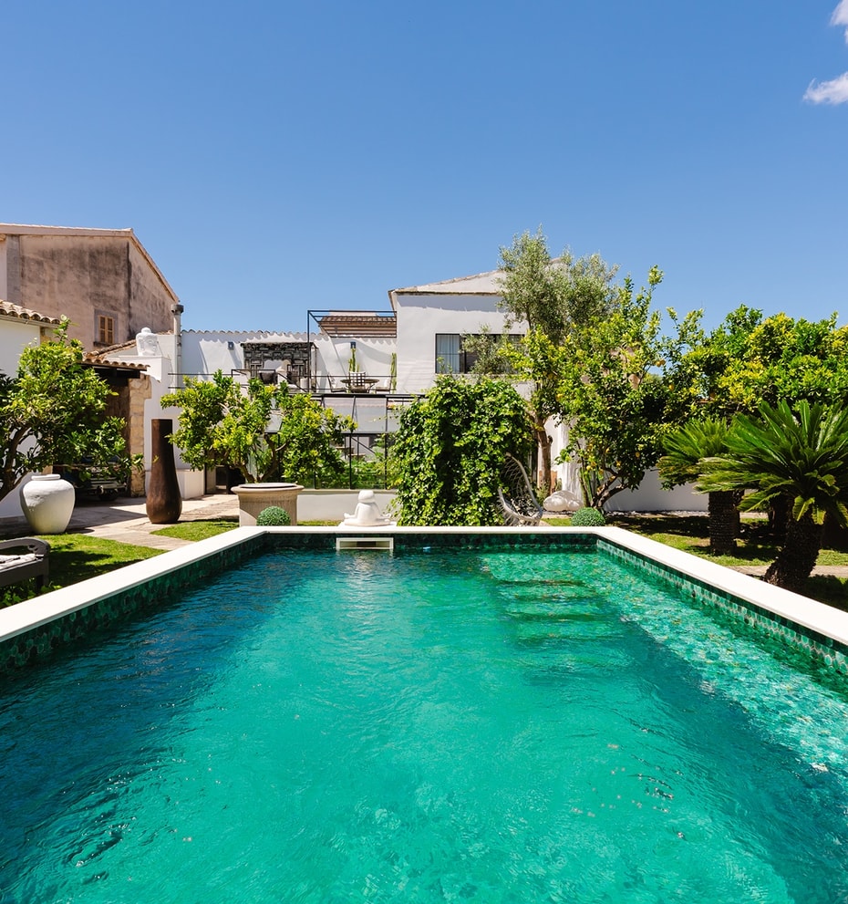 A rectangular swimming pool with turquoise mosaic tiles set within a lush, walled garden in Sencelles, featuring mature trees.