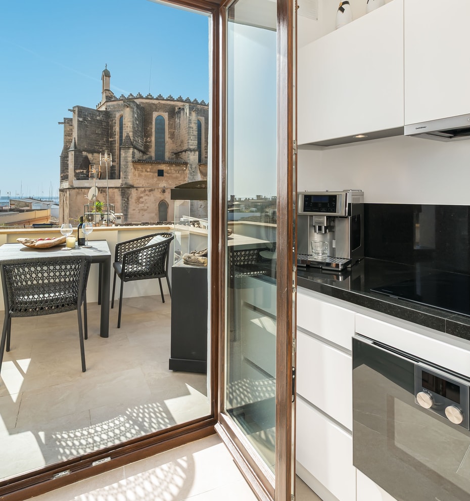 Modern white kitchen with black countertops opening to a sunny terrace with historic church views in Palma Old Town.