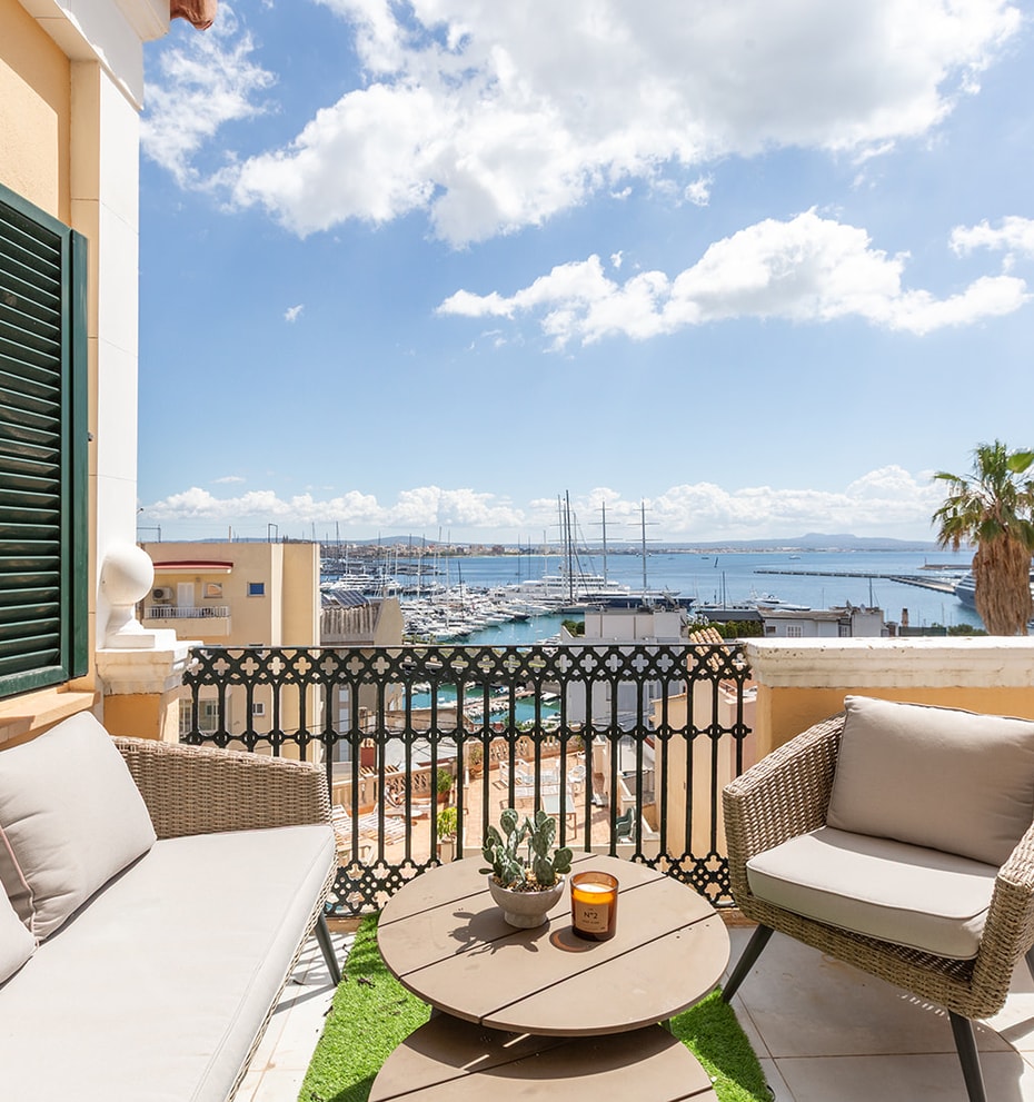 A sunny outdoor terrace featuring wicker seating, a small round table, and green shutters, overlooking a marina and sea.