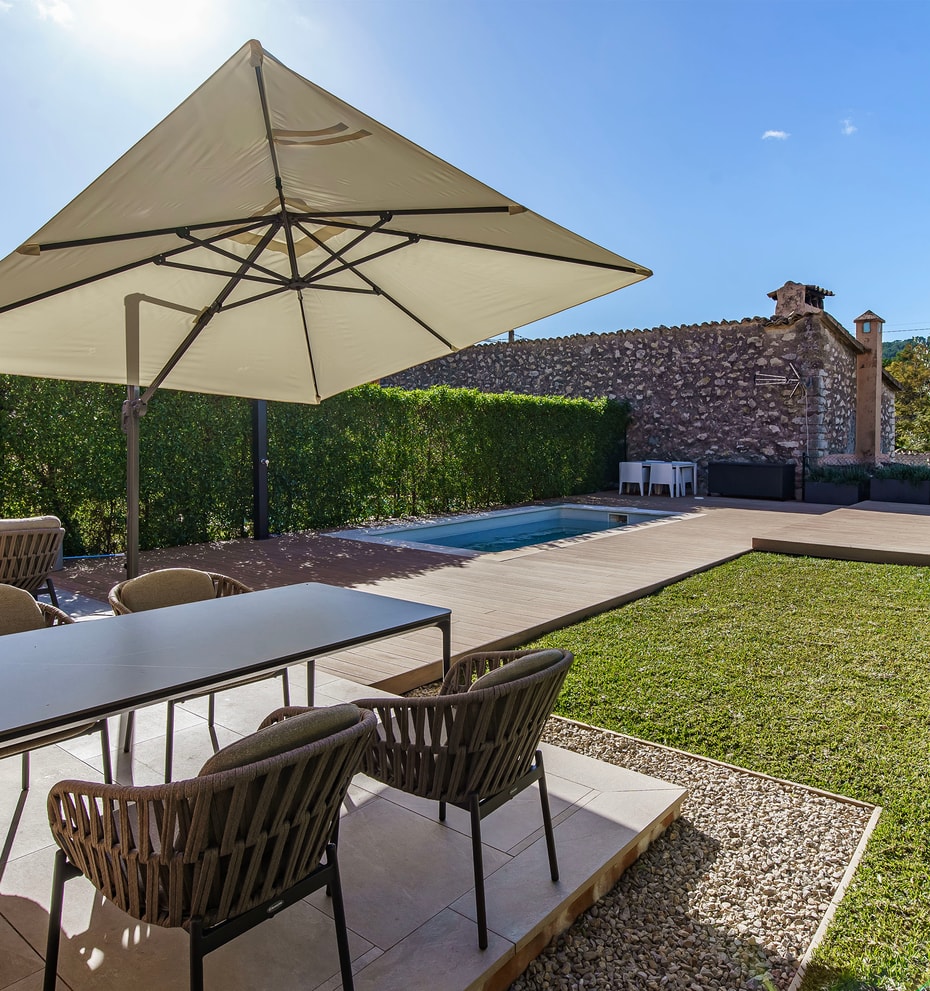 An outdoor dining terrace in Es Capdella featuring a rectangular table with woven chairs, a large beige parasol, and a small pool set against a natural stone wall.