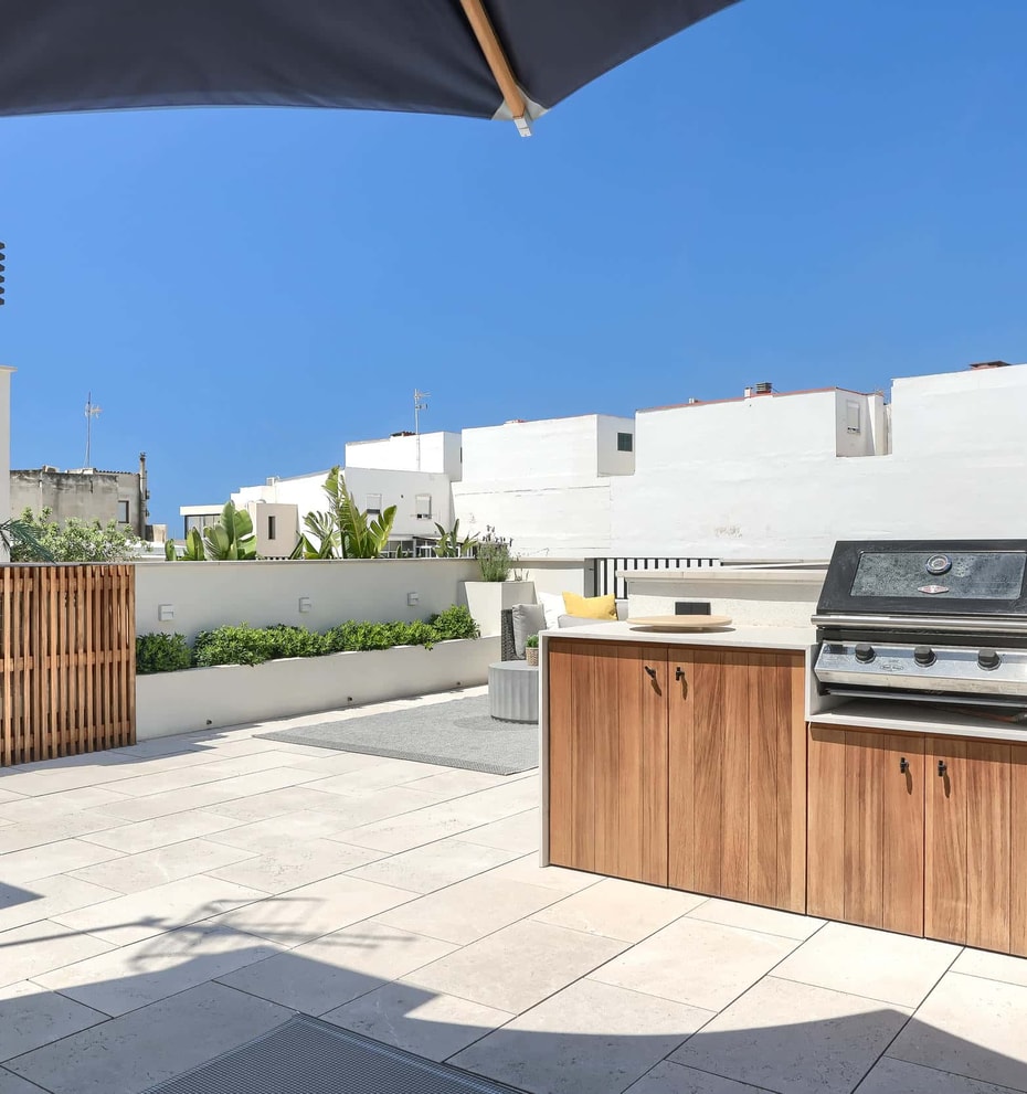 Rooftop terrace featuring a wooden outdoor kitchen cabinet with a built-in stainless steel grill, stone tile flooring, and slatted wood privacy fencing under a clear blue sky.