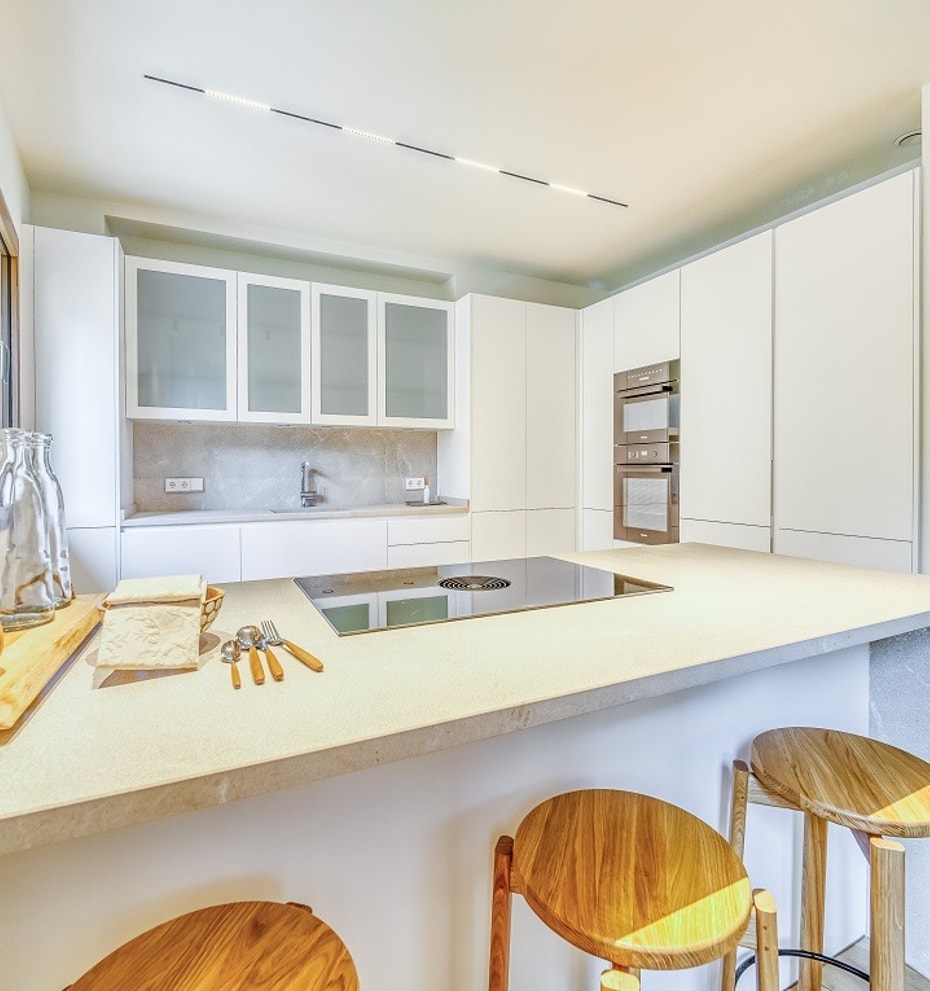 Modern kitchen with white cabinetry, integrated appliances, and a light stone island with wooden bar stools in the foreground.