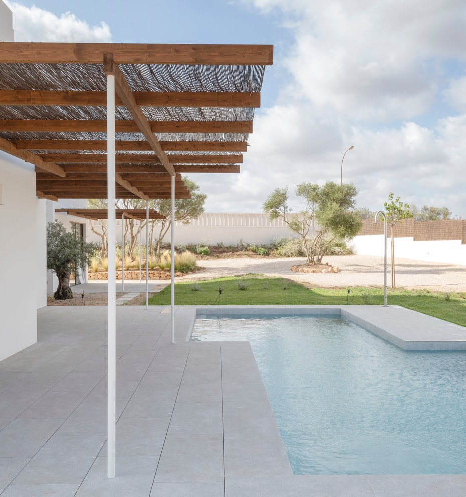 Modern pool terrace featuring white stone tiling and a timber pergola with reed shading next to a garden lawn.