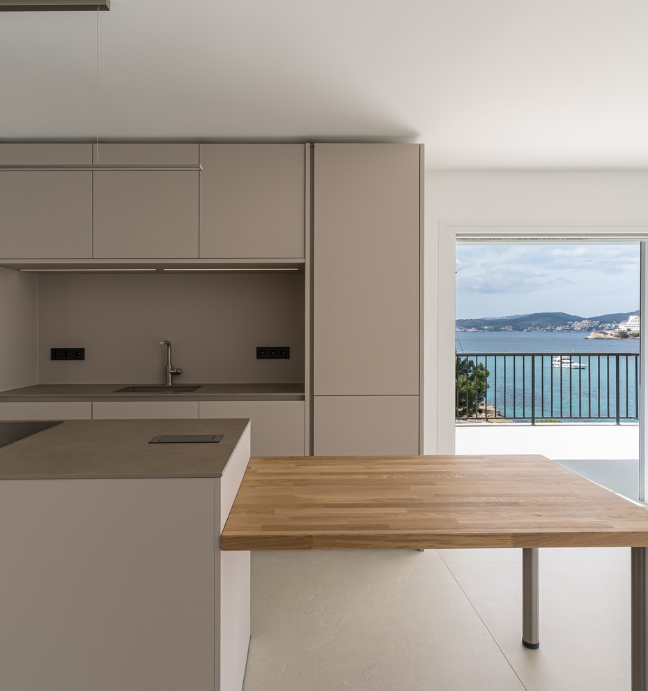 Minimalist kitchen with taupe cabinetry and an oak dining surface opening onto a sea-view terrace.
