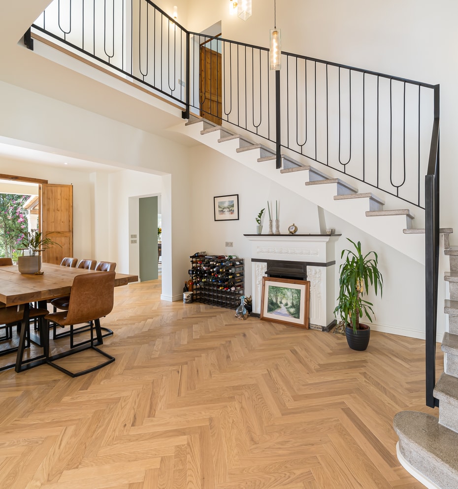 Sunlit living space with oak chevron floors, a black iron staircase, and a dining table near open wooden shutters.