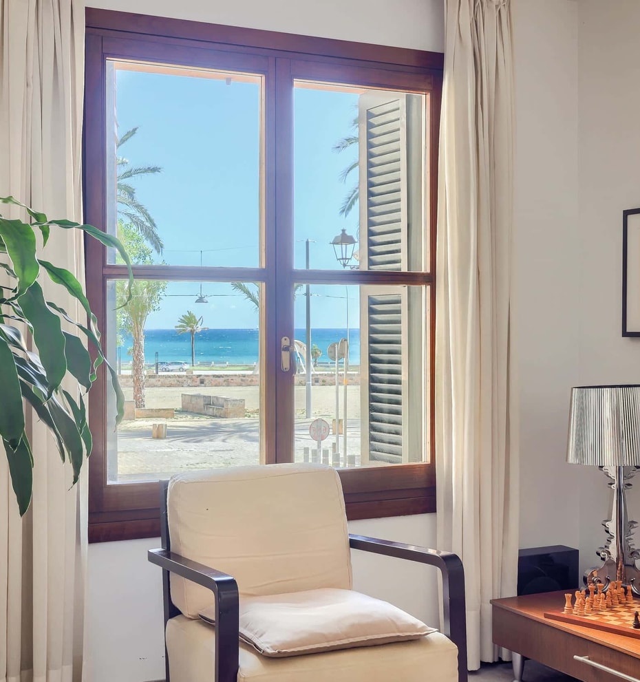A bright living space with a wooden window frame looking out to the sea and palm trees. An armchair is placed near the window next to a houseplant and a side table with a lamp.
