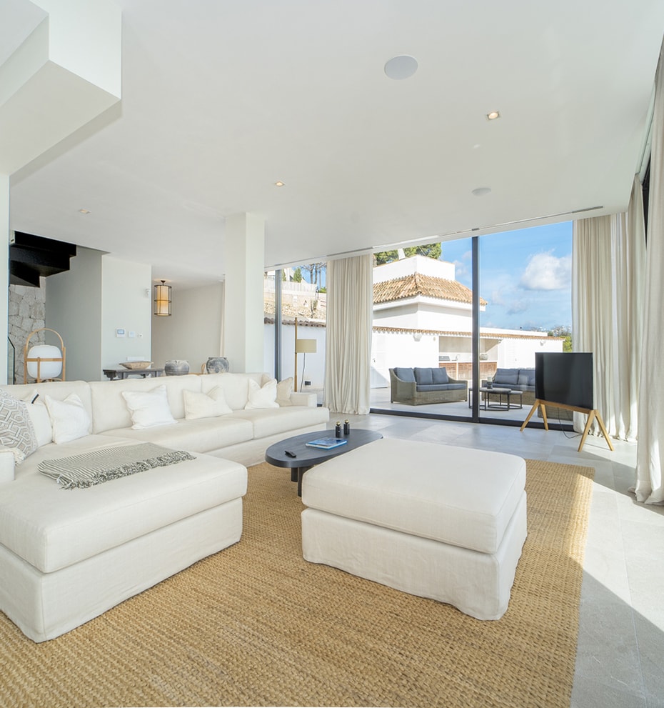 Bright living room with white L-shaped sofa, matching ottoman, woven area rug, and floor-to-ceiling glass sliding doors.